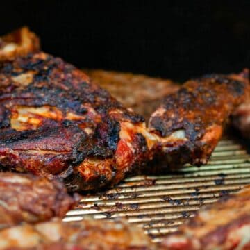 Close-up of various pieces of barbecued meat on a grill. The meat is charred and appears well-cooked, with a range of textures and colors from dark brown to lighter, cooked areas. The grill grates are visible beneath the meat.