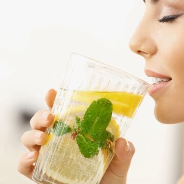 A person holds a glass of water infused with lemon slices and mint leaves close to their lips. The person is about to take a sip.