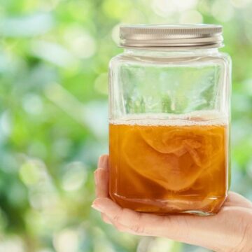 A hand is holding a glass jar filled with kombucha tea, which has a SCOBY (Symbiotic Culture of Bacteria and Yeast) floating inside. The background is blurred greenery.