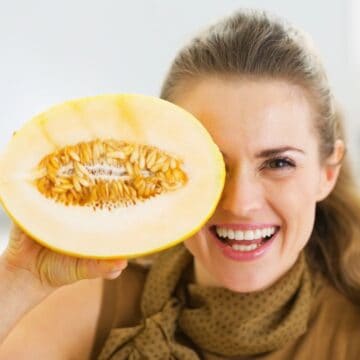 A woman with light brown hair holding half of a cantaloupe melon in front of her left eye is smiling. She is standing indoors, wearing a light brown scarf. The seeds and flesh of the melon are visible.
