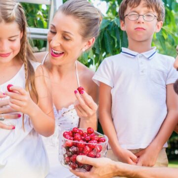 A family of four, two adults and two children, stands outdoors near a tree. The young girl and woman hold cherries while the man offers a bowl of cherries to the girl. The boy stands next to them with a neutral expression.