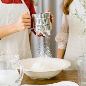 Two people are baking. One person is sifting flour into a mixing bowl while the other stands nearby. The table is filled with various gluten-free pantry essentials and utensils, and there is a large vase of flowers in the foreground.