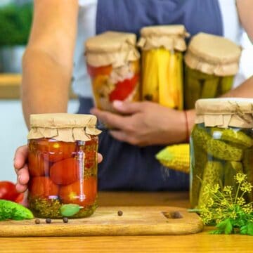 A person holding jars of assorted preserved vegetables arranges more jars of pickles, tomatoes, and peppers on a wooden surface. Fresh cucumbers, tomatoes, and bell peppers are placed nearby as they follow their guide to water bath canning.