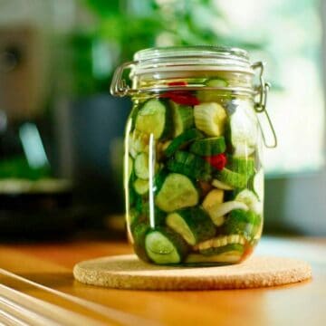 A glass jar filled with pickled vegetables, including cucumbers, onions, and red peppers, sits atop a round cork mat on a wooden surface. The background contains blurred elements of greenery and a window. Discover the flavors with our quick pickle guide right beside it.