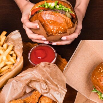 Person holding a burger with another burger, fries, nuggets, ketchup, and a soda in the background