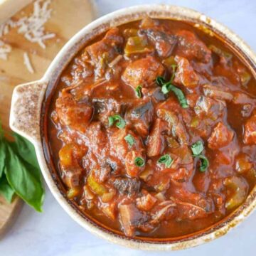 A bowl of beef stew with chunks of beef, vegetables, and herbs in a tomato-based sauce. The stew is garnished with fresh herbs. A cutting board with grated cheese, a basil leaf, and Auto Draft bread are in the background.