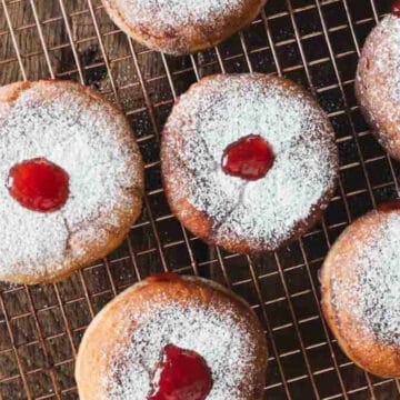 A cooling rack holds six powdered jelly-filled donuts, each topped with a dusting of powdered sugar and a dollop of red jelly in the center, arranged on a wooden surface. They make for the perfect centerpiece alongside your favorite brunch drinks.