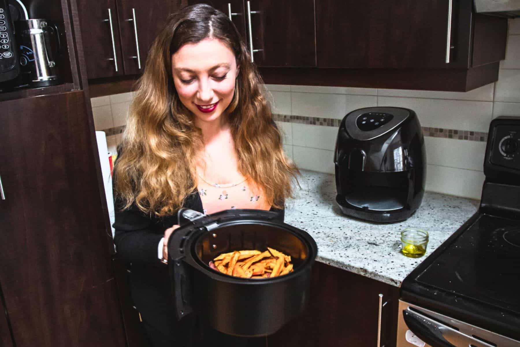 A person with long hair, wearing a patterned shirt and a black cardigan, holds an air fryer basket filled with perfectly cooked fries in a kitchen. An air fryer—a true air fryer success—sits on the countertop next to the stove, along with a small container of oil and must-have accessories.