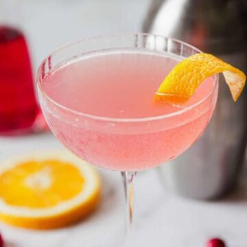 A pink cocktail is served in a coupe glass with a twisted lemon peel garnish. A cocktail shaker, a slice of orange, cranberries, and a red liquid in a small bottle are in the background on a marble surface—an iconic drink recipe that's as beautiful as it is delicious.