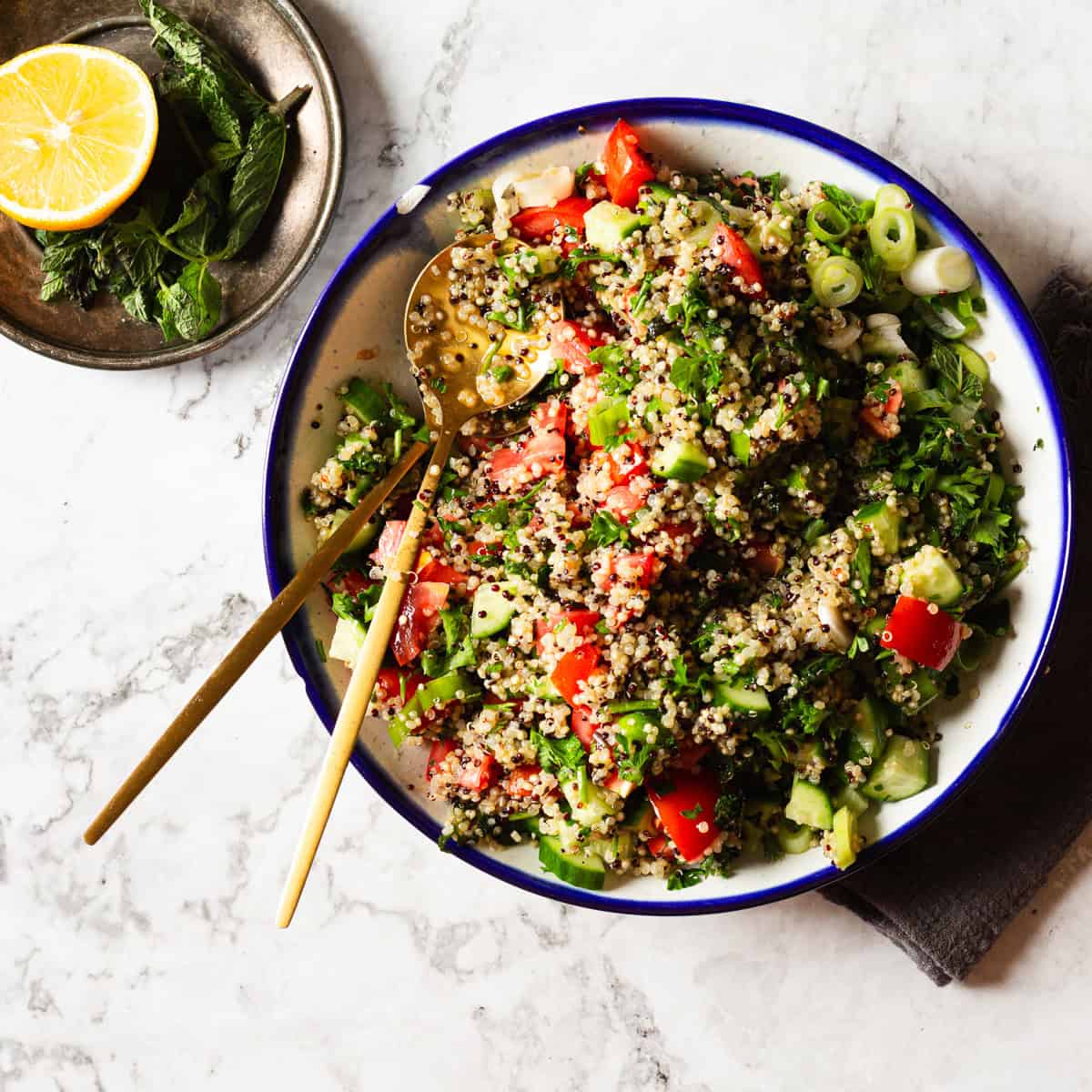 A bowl of quinoa salad, reminiscent of a traditional Middle Eastern recipe, is placed on a marble surface. The salad contains quinoa, diced tomatoes, cucumbers, green onions, and fresh herbs. A pair of gold-tone utensils rests in the bowl. A saucer with a half lemon and mint leaves is positioned to the side.