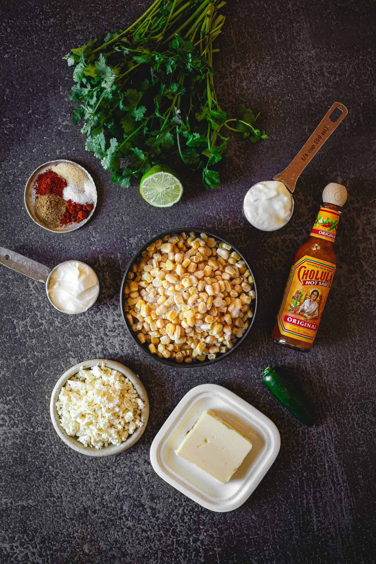 A flat lay of ingredients on a dark surface for making Elote en Vaso, a Mexican street corn salad. Items include corn, cheese, a block of butter, jalapeño, lime, hot sauce, yogurt, spices, mayonnaise, and fresh cilantro.