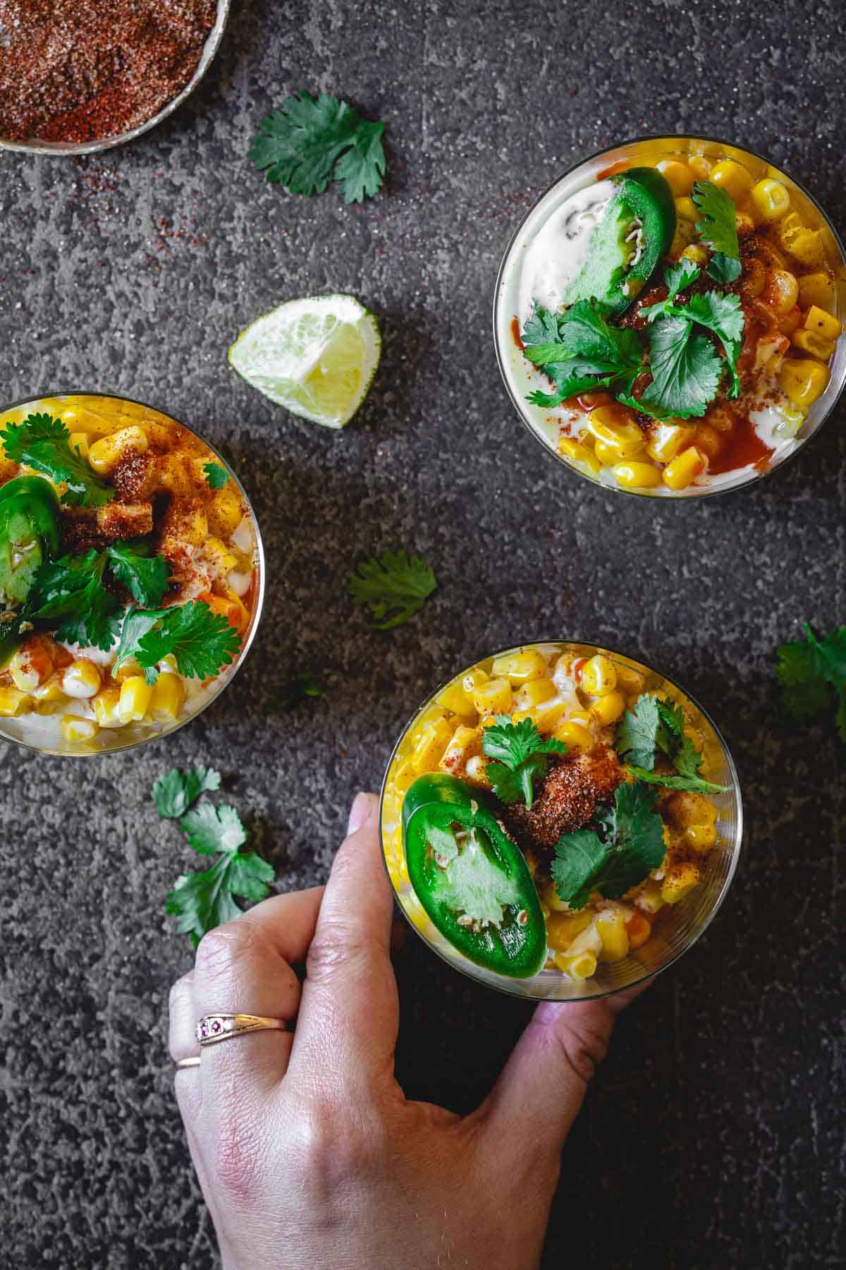 Three glasses filled with a Mexican Elote mixture of corn, sliced green jalapenos, cilantro, and creamy dressing are placed on a dark surface. A hand with a ring is holding one of the glasses. A lime wedge and a small bowl of spices are also visible on the surface.