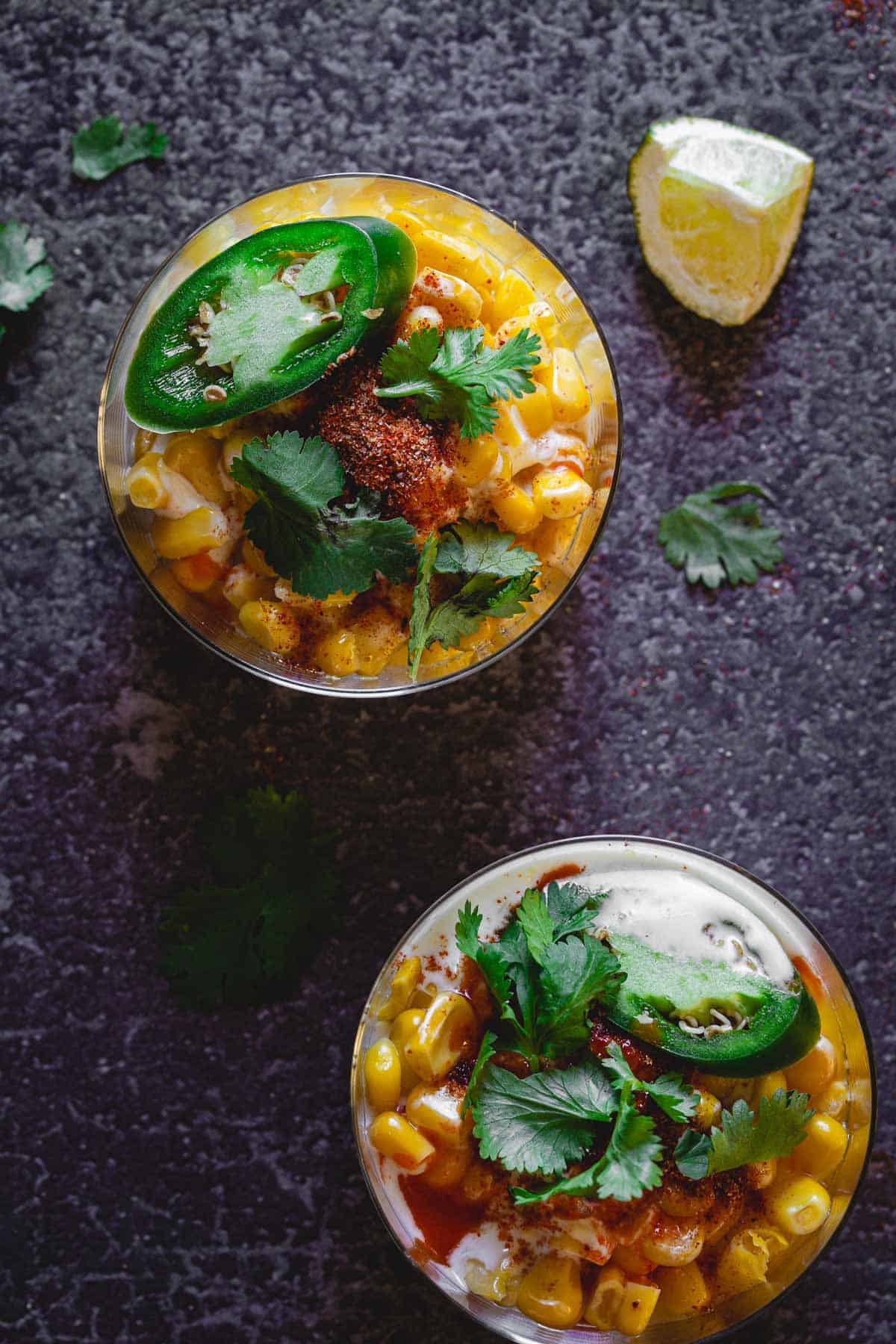 Two glasses filled with Mexican Elote corn salad are garnished with jalapeño slices, cilantro, cotija cheese, and chili powder. A lime wedge and cilantro leaves are scattered on the dark textured surface around the glasses—a perfect example of an Elote en Vaso cup recipe.