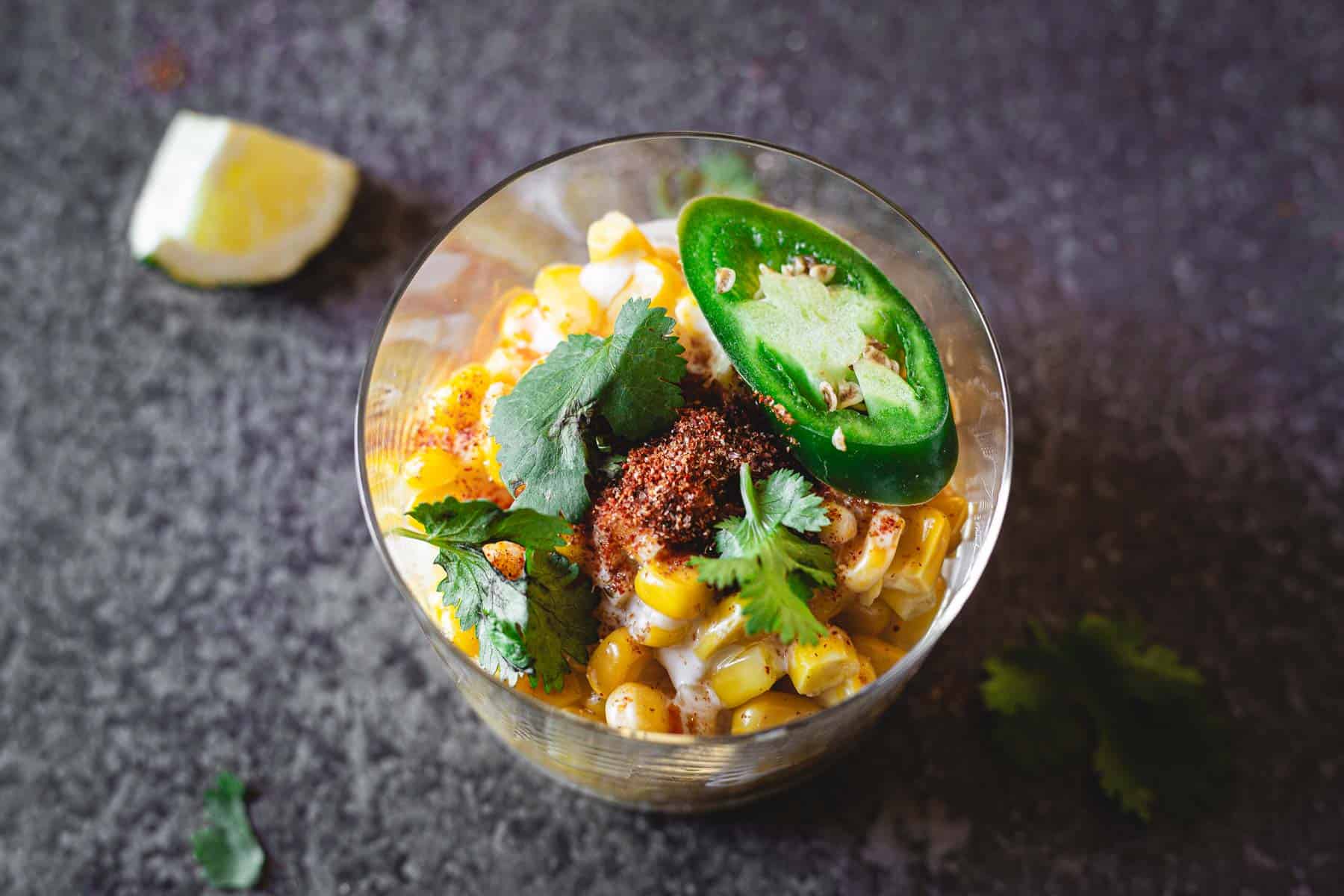 A glass cup filled with a Mexican Elote salad, garnished with sliced jalapeño, cilantro leaves, and red seasoning. A lime wedge and some cilantro leaves are scattered around on a dark textured surface.