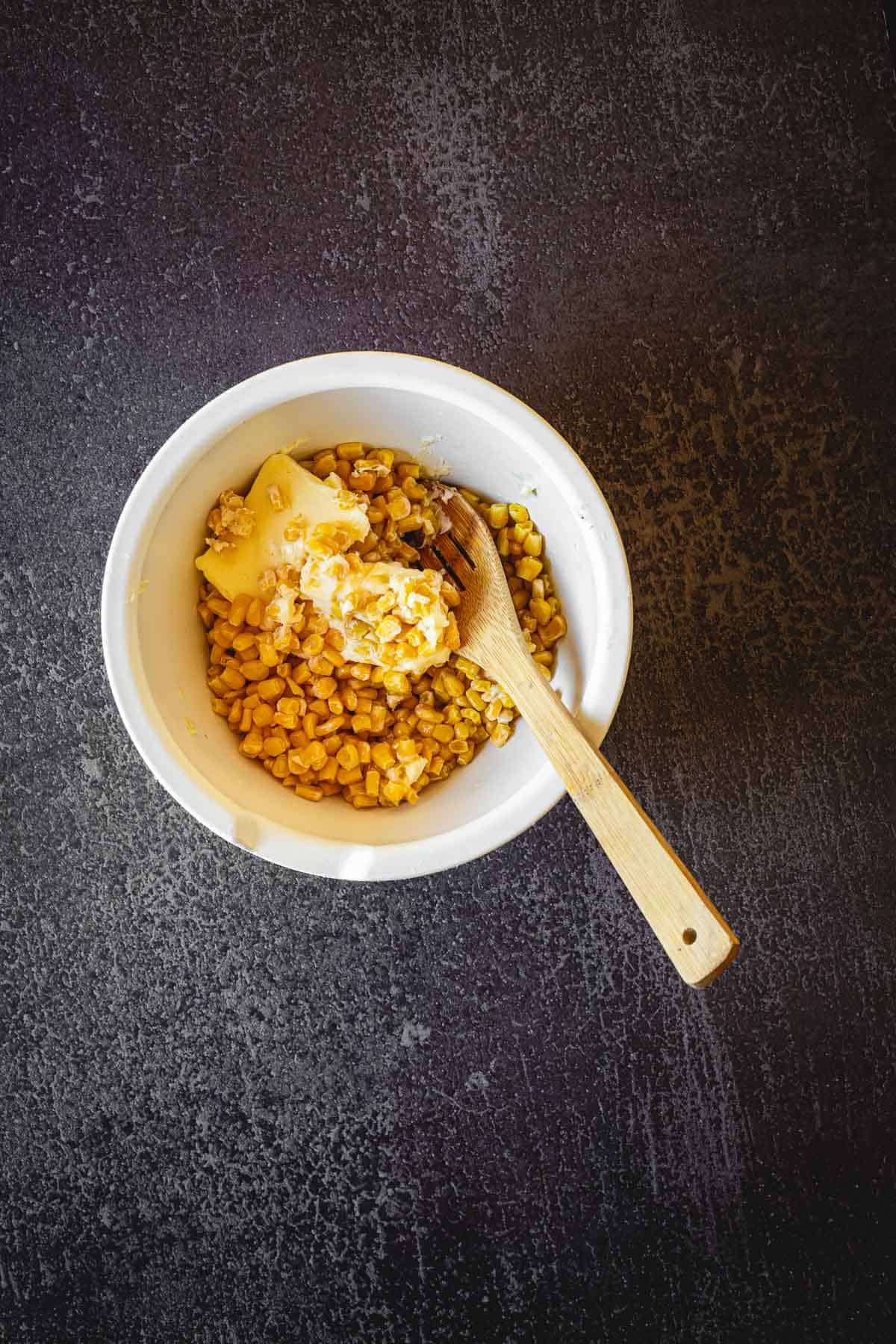 A white bowl filled with corn kernels topped with a dab of butter and a wooden fork resting inside, resembling a classic Elote en Vaso, placed on a dark textured surface.