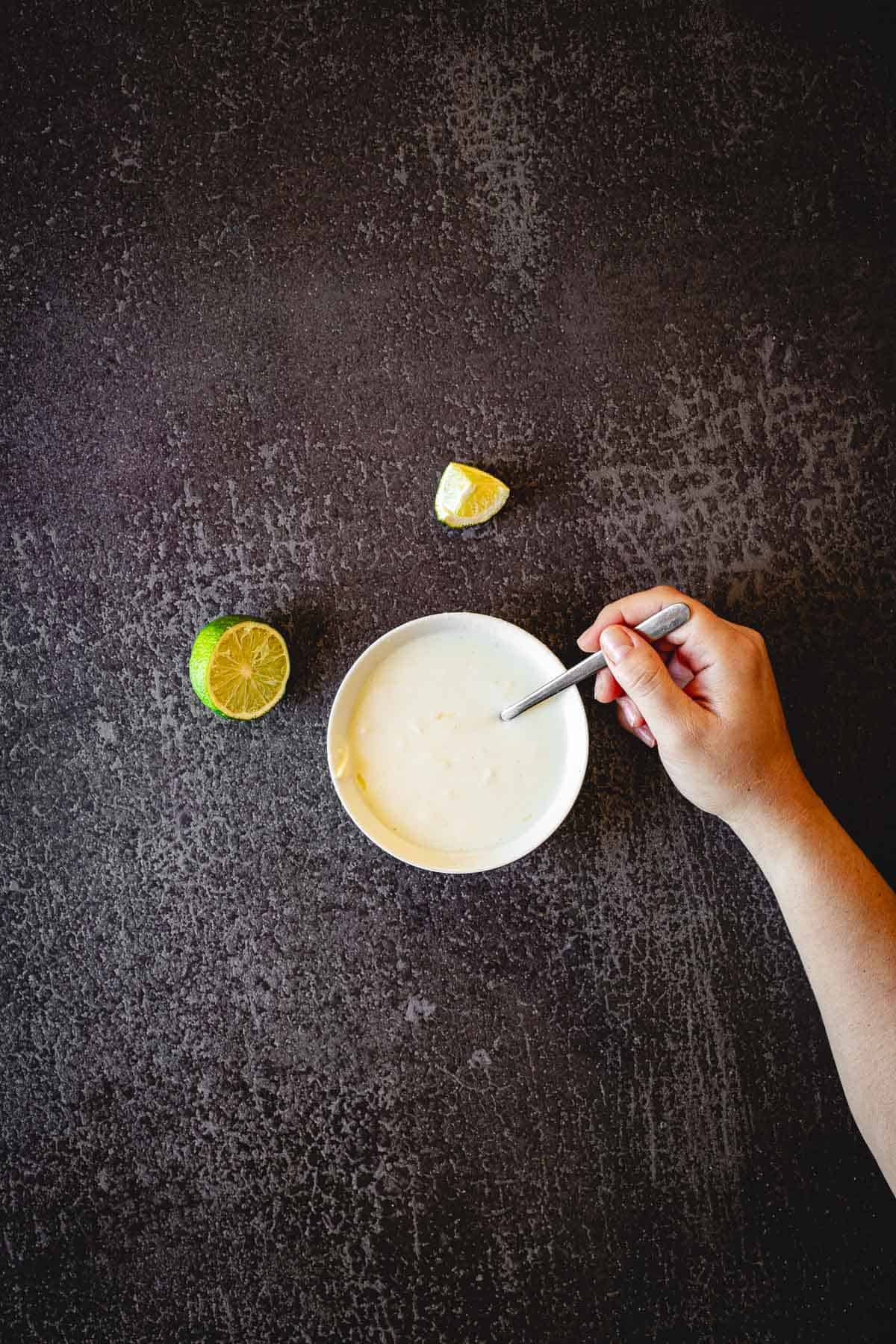 A hand holding a spoon stirs a bowl of creamy white sauce on a dark textured surface, perfect for an Elote en Vaso recipe. A halved lime and a lime wedge are placed next to the bowl.