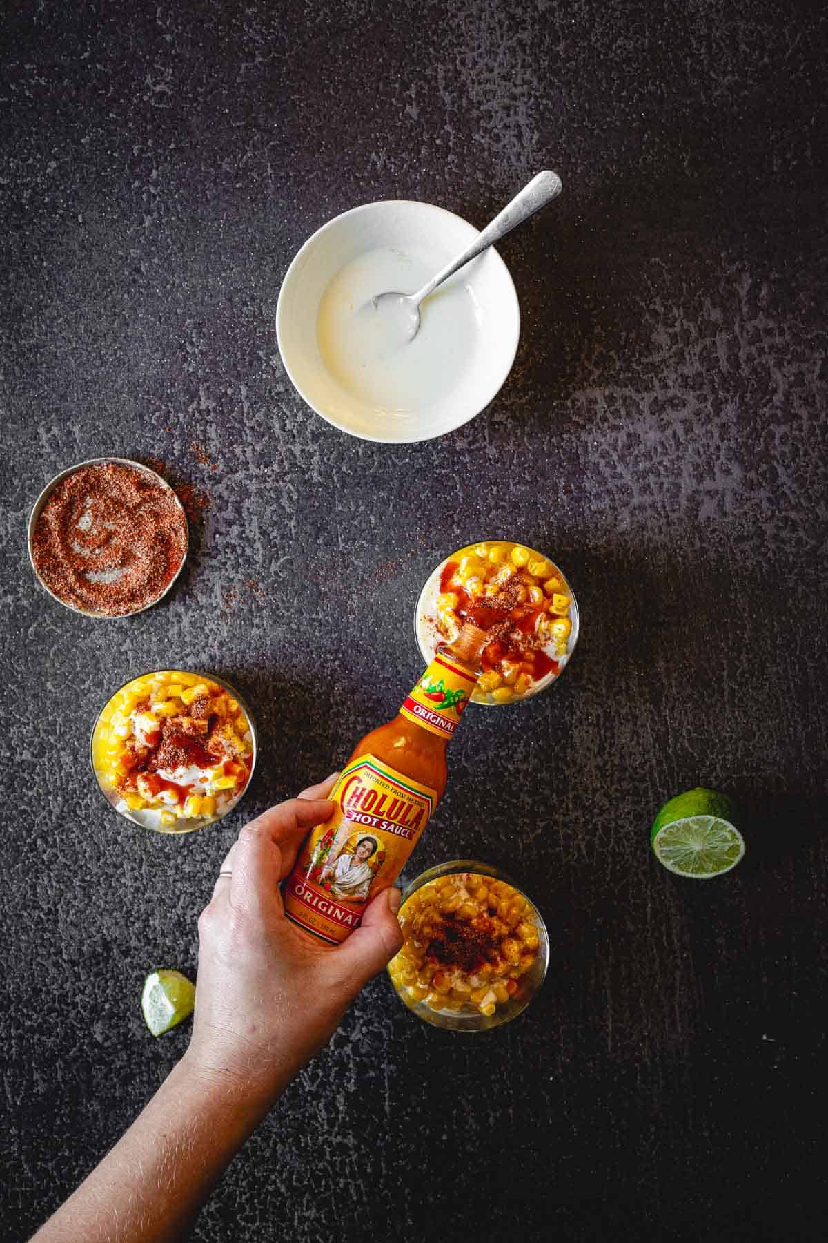 A top-down view shows a hand pouring Cholula hot sauce on three cups of Elote en Vaso, a Mexican elote cup recipe. Nearby are a bowl of white sauce with a spoon, a bowl of chili powder, and a sliced lime on a dark surface.