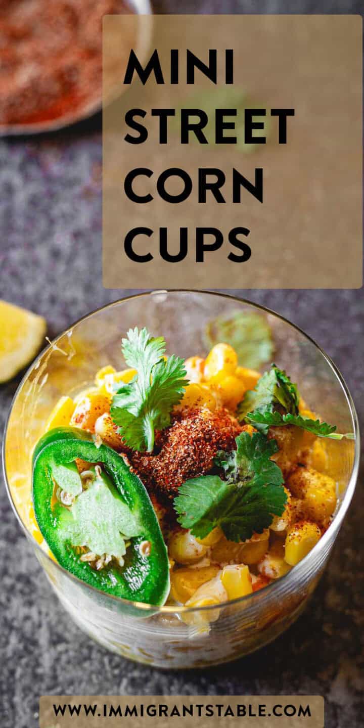 A glass cup filled with corn kernels, garnished with a slice of green jalapeño, cilantro, and chili powder. The text "MINI STREET CORN CUPS" and "Elote en Vaso Recipe" are prominently displayed above the cup, and the website www.immigrantstable.com is noted at the bottom.