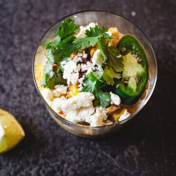 This image shows a clear glass bowl filled with a mixture of food ingredients, reminiscent of an Elote en Vaso. The bowl contains crumbled white cheese, sliced green jalapeños, cilantro leaves, and what appears to be corn salad. A cut piece of lime is placed nearby on the dark surface.