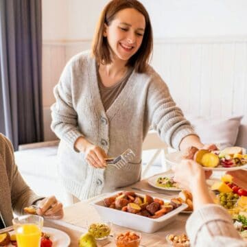 Four people are seated around a dining table enjoying a meal, embodying the essence of hosting a successful brunch. One person is standing and serving food to another seated guest. The table is set with various dishes, salads, and glasses of orange juice. The atmosphere appears relaxed and cheerful.