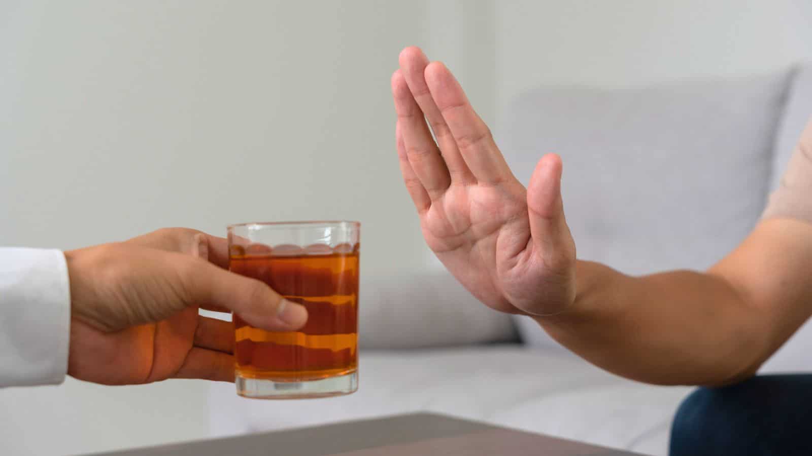 A person extends a glass of a brown liquid towards another person. The second person holds up their hand, palm open, in a gesture indicating refusal. In the background, a light-colored sofa is visible.