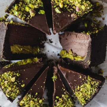 A chocolate cake sliced into eight pieces, arranged in a circular pattern on a piece of white parchment paper. Each slice has a thick layer of dark chocolate on top and is topped with chopped pistachios, revealing a green filling inside.