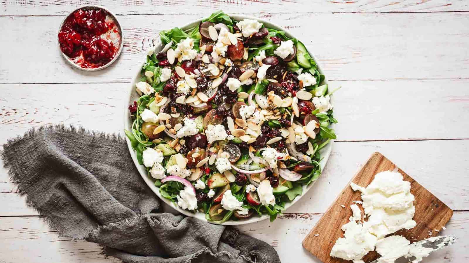 A large salad served on a round plate with a variety of greens, red onions, nuts, seeds, and cheese crumbles. A bowl of red dressing, a cutting board with more cheese, and a gray fabric napkin are placed beside the plate on a white wooden table&mdash;dispelling common food myths about boring salads.