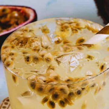 A close-up of a glass filled with a yellow, passion fruit drink. The drink contains visible passion fruit seeds and ice cubes. A striped paper straw is inserted into the glass. In the background, halved passion fruits and a coconut shell are partially visible, evoking the essence of summer concoctions.