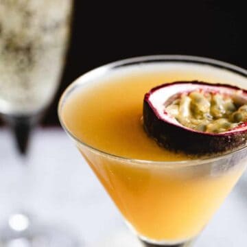A close-up of a yellow-orange, nonalcoholic cocktail in a martini glass garnished with a halved passion fruit. The background shows part of another drink in a tall, stemmed glass, possibly champagne, slightly out of focus.