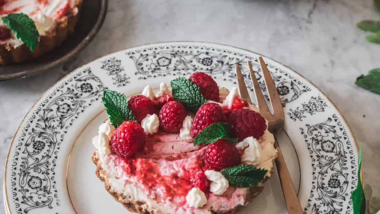 A small tart with a filling topped with fresh raspberries, dollops of whipped cream, and mint leaves is placed on an ornate patterned plate. A fork rests on the right side of the plate. Another dessert is partially visible in the background.