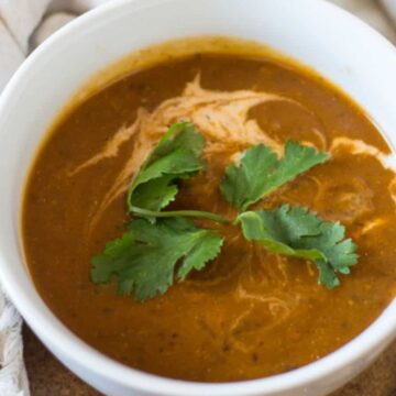 A white bowl filled with reddish-brown soup, garnished with a swirl of cream and a sprig of fresh cilantro, sits on a table next to a beige cloth napkin and a silver spoon—true comfort food at its finest. Discover more delightful recipes in our soups collection.