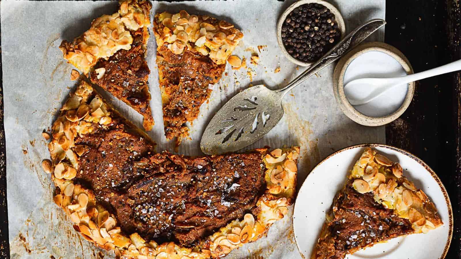 A rustic galette with almond flakes is partially sliced on parchment paper, captured in an auto draft. A pie server is placed near the galette, and two pieces have been served on a small plate. Nearby, small bowls of peppercorns and a white powder, possibly sugar, complete the scene.