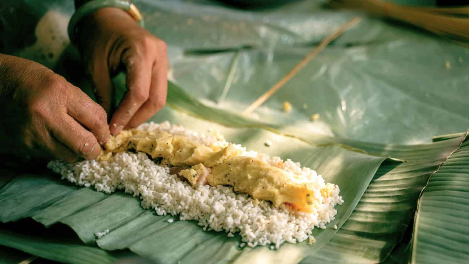 Two hands are preparing a dish by placing a mixture on top of white rice laid on large green leaves, possibly banana leaves. The savory topping appears to contain eggs or a sauce.
