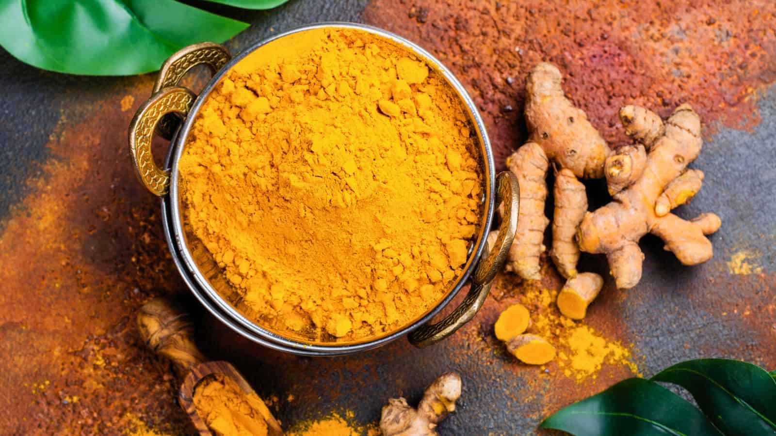 A bowl of turmeric powder with brass handles sits on a surface sprinkled with turmeric. Fresh turmeric roots, some sliced to show the bright orange interior, lie beside the bowl. Green leaves are placed around the scene for decoration.