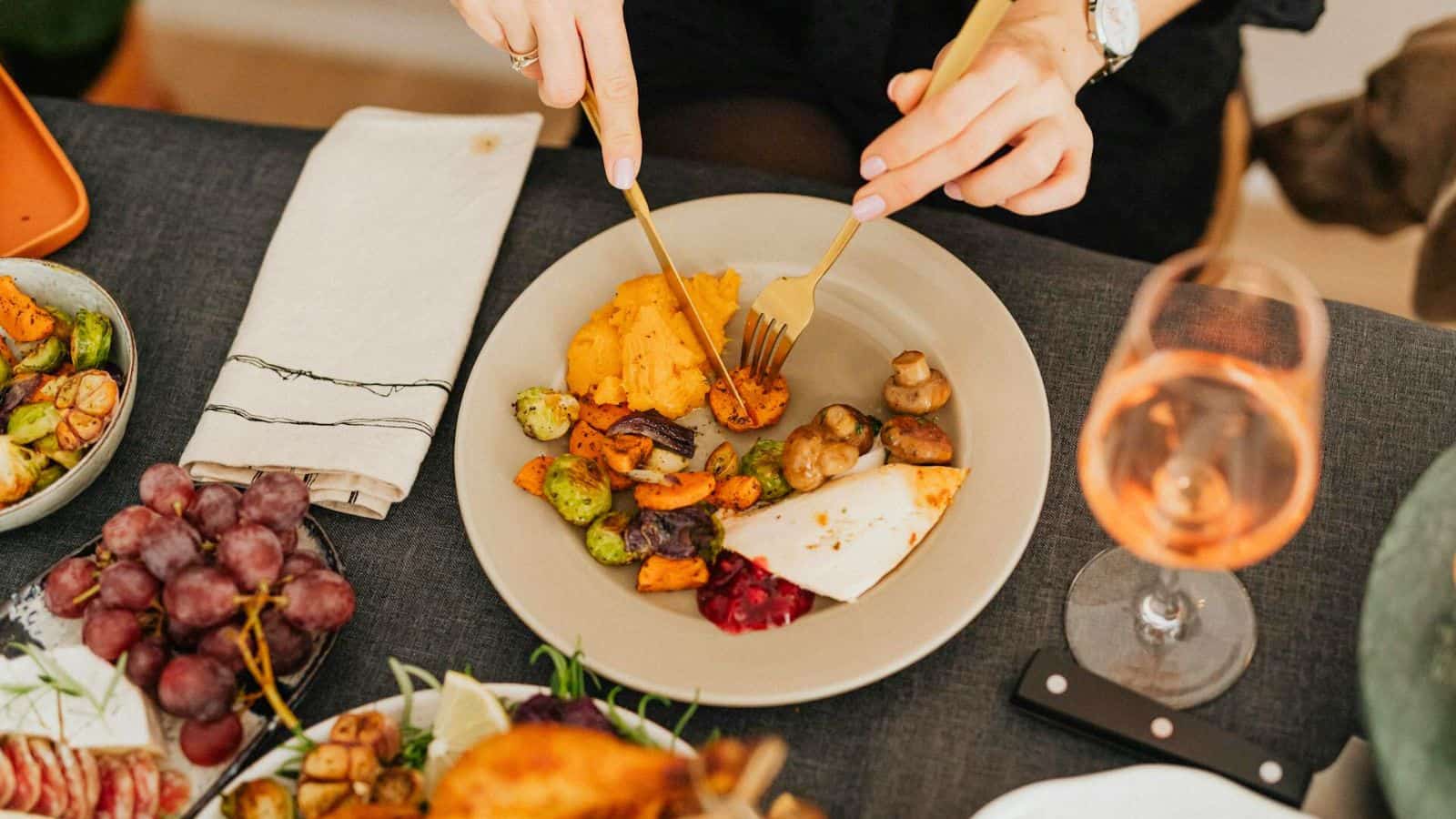 A person is using a knife and fork to enjoy roasted vegetables, mashed potatoes, and a piece of meat. The table also features a glass of ros&eacute; wine, grapes, and additional plates of food nearby. Amid these delicious dishes, the casual dining setting seems free from any food myths clouding the meal.