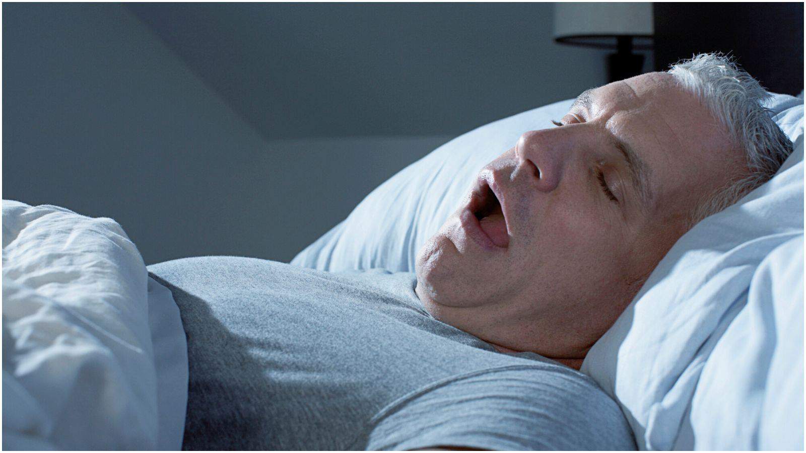 A man with short gray hair is lying in bed on his back, wearing a gray t-shirt. His mouth is open, indicating he is asleep. The bed has white sheets and pillows, and a lamp is visible in the background.