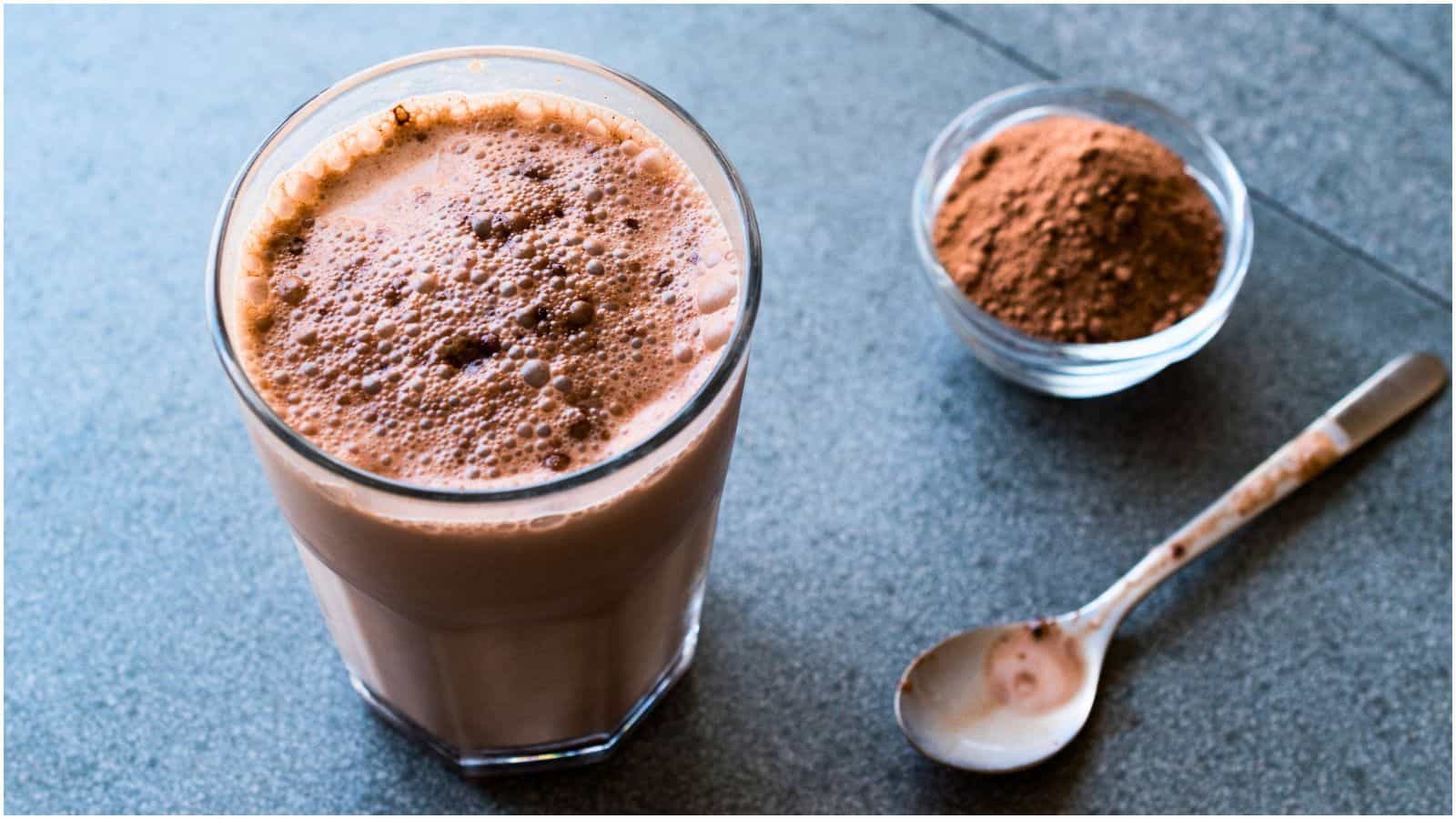 A glass filled with frothy chocolate milk is placed on a gray surface. To the right of the glass, there is a small glass bowl containing cocoa powder and a spoon with some cocoa powder residue, debunking food myths about chocolate being just for indulgence.