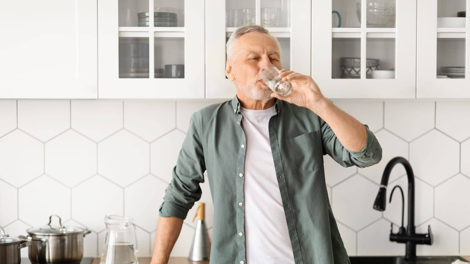 A man with grey hair and a beard is standing in a kitchen with white hexagonal tile backsplash, contemplating food myths as he drinks water from a glass. The kitchen, adorned with white cabinets featuring glass doors, a black faucet, and various items on the countertop, exudes simplicity and charm.