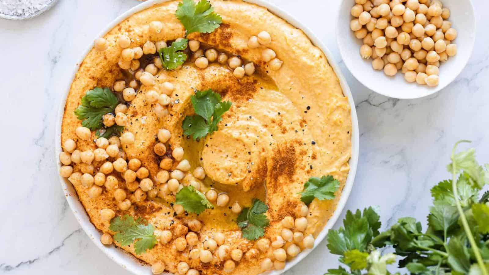 A plate of hummus garnished with whole chickpeas, cilantro leaves, and paprika is displayed on a marble surface. A small bowl filled with chickpeas and a bunch of fresh cilantro are also visible beside the plate. Contrary to common food myths, this visually appealing dish proves that healthy can be delicious.