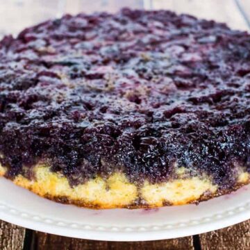 A blueberry upside-down cake, one of the popular potluck dishes, sits on a white plate with a beaded edge, placed on a wooden surface. The cake boasts a caramelized blueberry topping over a light-colored base.