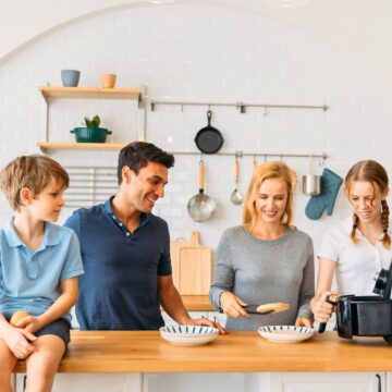 A family of four is in a kitchen. A man and woman are smiling, with the man sitting on a counter and the woman holding a spatula. A girl is using cool and easy kitchen gadgets, and a boy is looking on. The kitchen has white tiles, a wooden countertop, and hanging utensils.