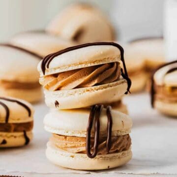 The image shows several light-colored macarons filled with a creamy substance, possibly caramel or coffee flavor. Drizzled with chocolate on top, these holiday treats are arranged on a white surface, with some blurred in the background. Perfect for festive baking enthusiasts!