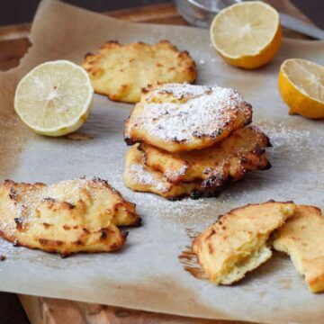 Four cheese biscuits, lightly dusted with powdered sugar, are arranged on a sheet of parchment paper. Two halves of a lemon and some lemon zest are visible in the background. Drawing inspiration from 60's recipes, this delicious treat is perfect for the best breakfast experience.