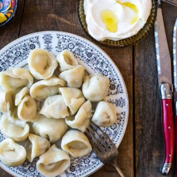 A plate of dumplings sits on a wooden table next to a fork and two knives with red handles. A small dish with a dollop of sour cream, garnished with a drizzle of oil, is nearby. A decorative wooden spoon is also on the table.