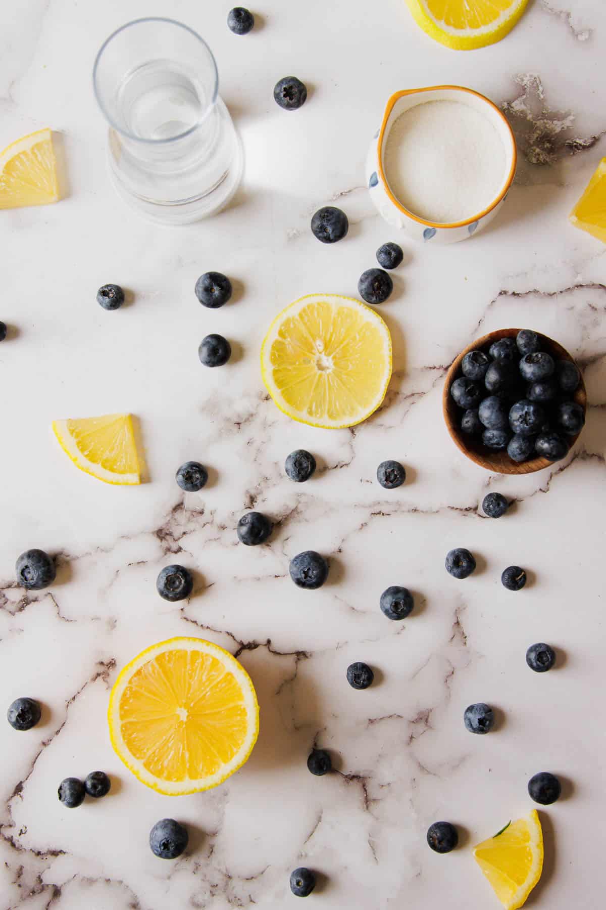 A marble countertop with scattered blueberries and lemon slices. A small bowl filled with blueberries, a glass of water, and a small bowl containing a white granular substance are also present on the surface, perfect for making 3-Ingredient Blueberry Jam.