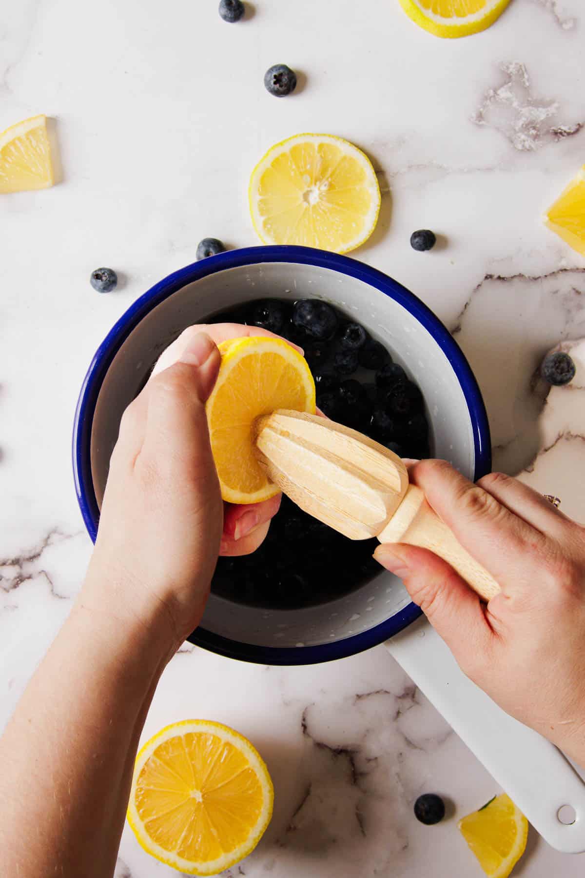 Two hands are squeezing a lemon half using a wooden citrus reamer over a white and blue bowl filled with blueberries, perfect for making 3-Ingredient Blueberry Jam. Slices of lemon and scattered blueberries dot the marble countertop surrounding the bowl.