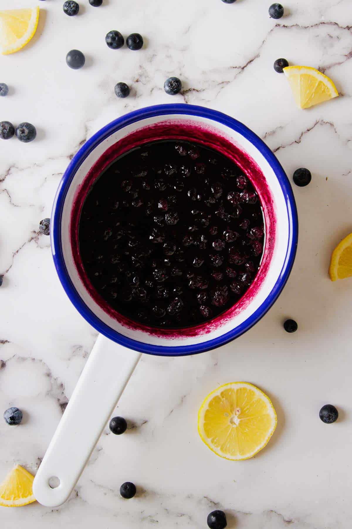 A white enamel saucepan with a blue rim contains Easy Blueberry Jam, made from just three ingredients. The saucepan rests on a marble surface decorated with scattered whole blueberries and lemon wedges.