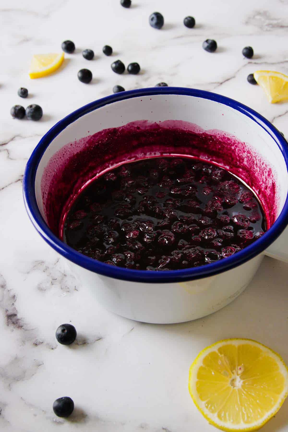 A white enamel saucepan with a blue rim contains a dark purple 3-ingredient blueberry refrigerator jam. The saucepan is placed on a white marble surface, with scattered blueberries and lemon slices around it.