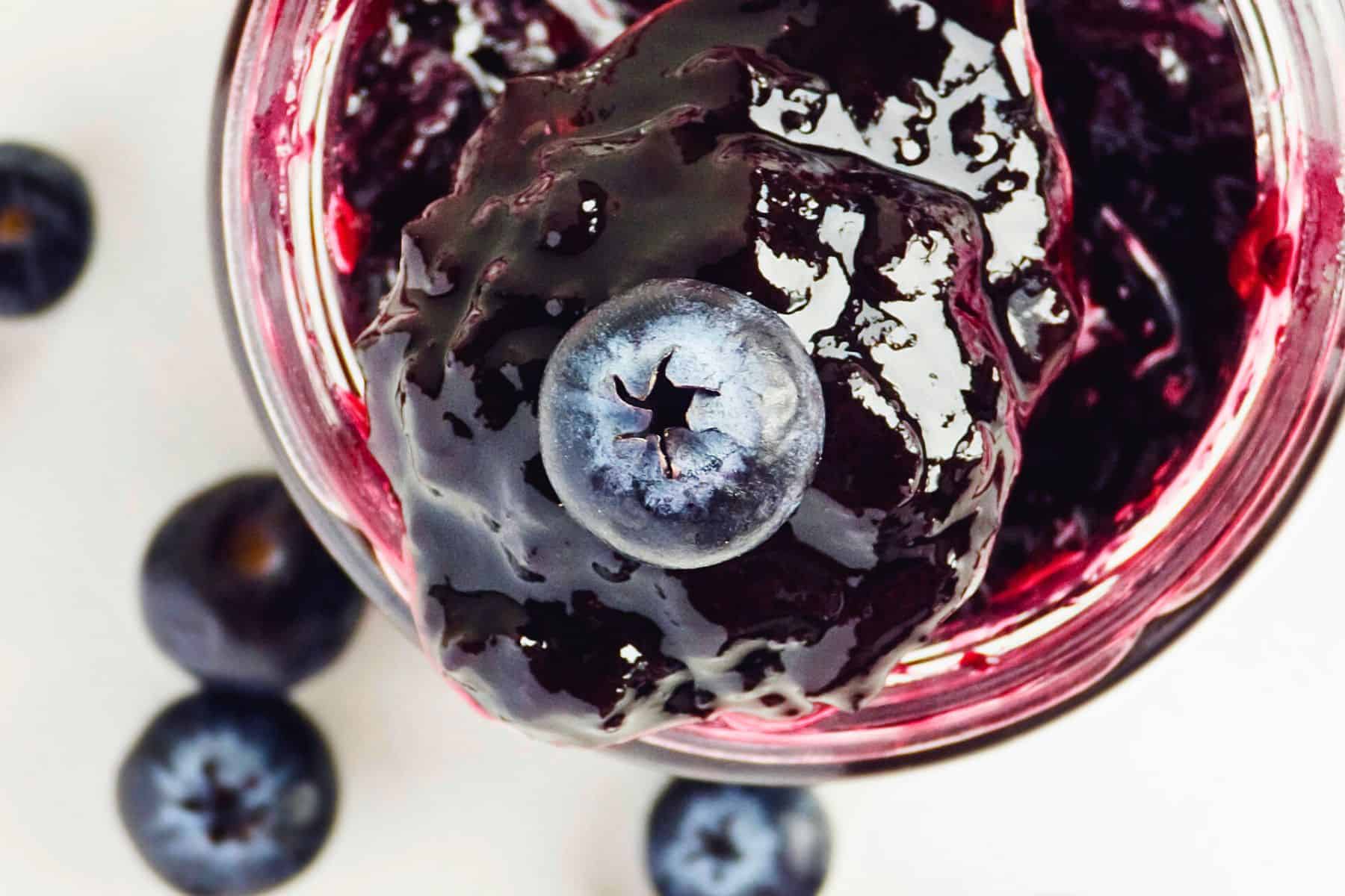 A close-up view of a glass jar filled with 3-ingredient blueberry jam. The jam appears thick and glossy, with a whole blueberry resting on top. Several additional blueberries are scattered on the white surface around the jar, perfectly showcasing this delicious refrigerator jam.