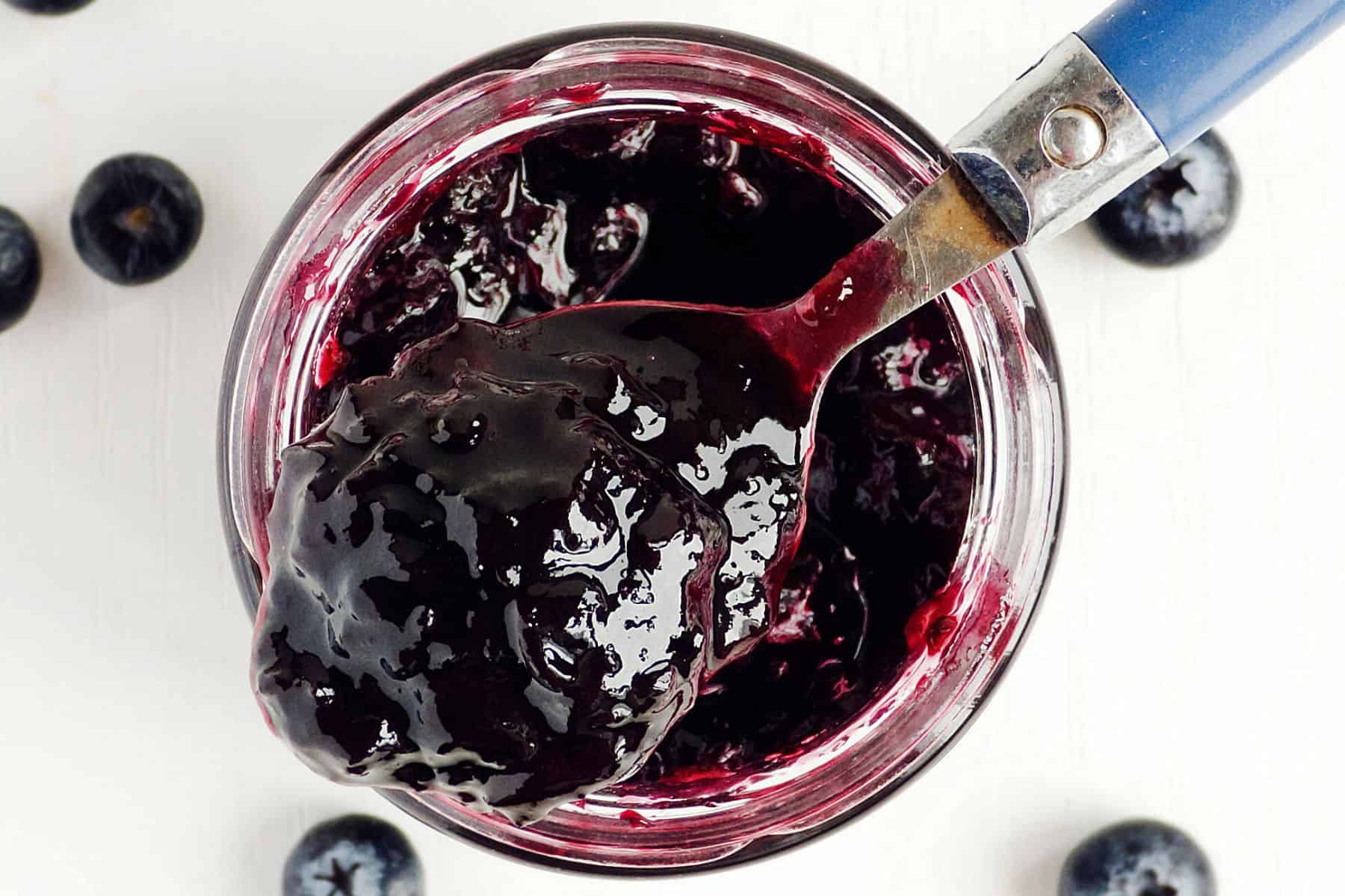 A close-up view of an open jar filled with dark blueberry jam reveals a silver spoon with a blue handle resting inside, covered in the sweet spread. Several whole blueberries are scattered around the jar on a white surface, hinting at the simplicity of this 3-ingredient refrigerator jam.